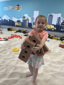 A small girl laughing and smiling while surrounded by construction toys in the Baha Play Sand pit at the Top children's amusement center.