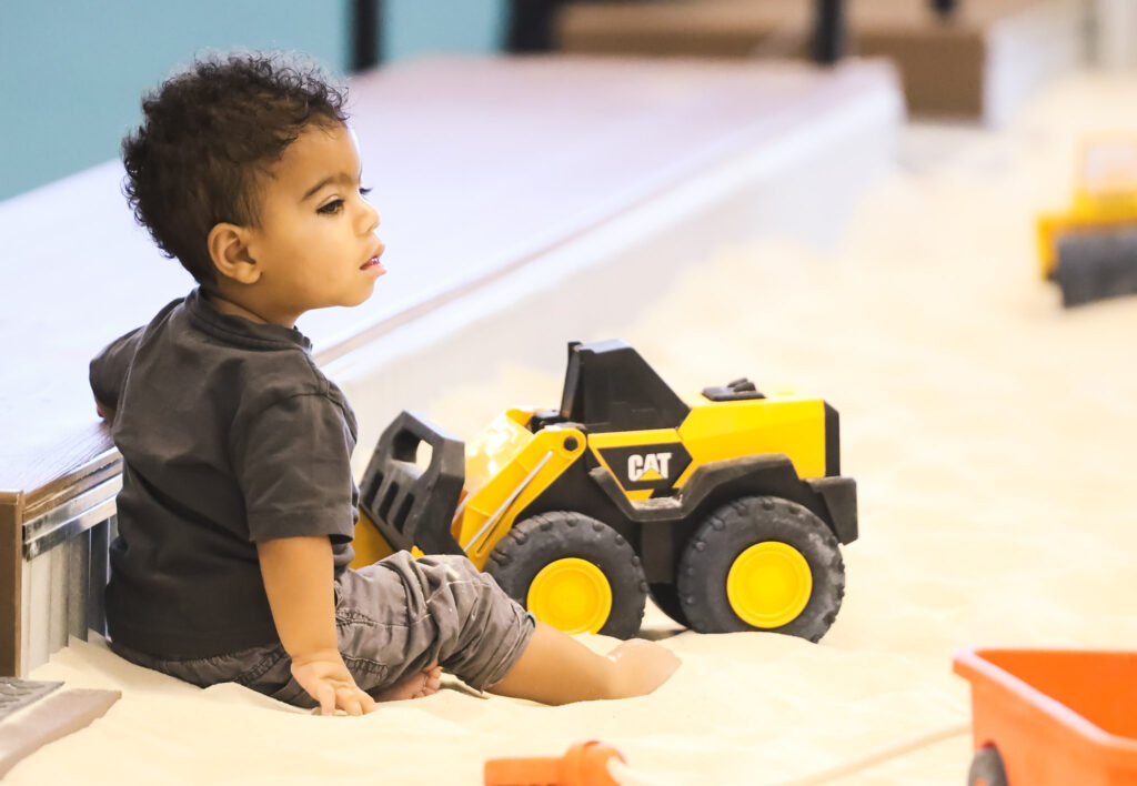 Young boy pushing a toy construction truck in an indoor sandbox playground.