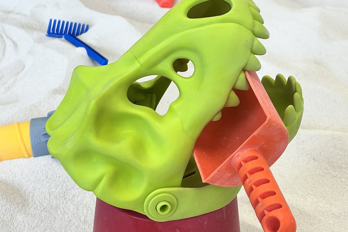 A bright green dinosaur head sand toy with a scoop in its mouth, sitting in the sandbox at The Sand Place in Aurora, Colorado