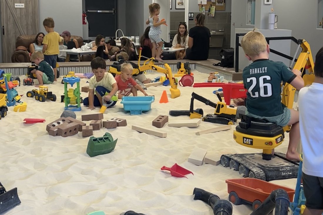 Children building with trucks, bricks and a ride-on CAT excavator in the big sandbox at The Sand Place in Aurora, Colorado, while parents sit at tables behind them