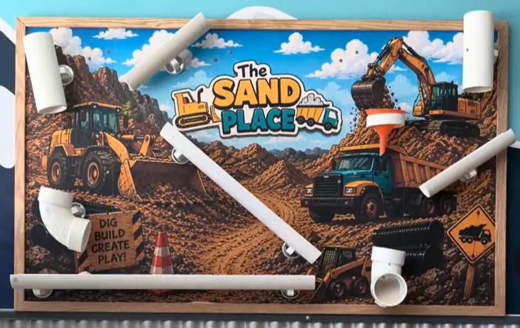 The new magnetic sand wall at The Sand Place, a framed construction-scene mural with white tubes and a funnel arranged on it so kids can build paths for the sand to flow through