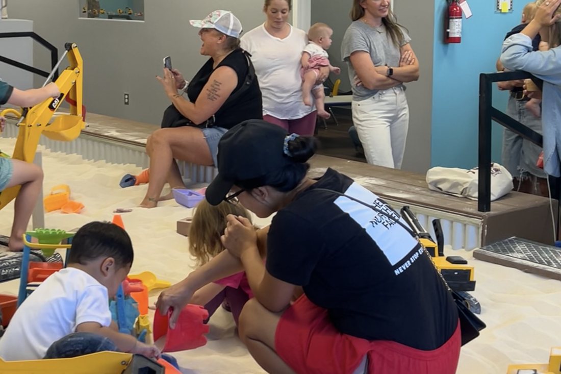 A parent kneeling in the sand helping two young children fill a bucket during a preschool playdate fundraiser at The Sand Place